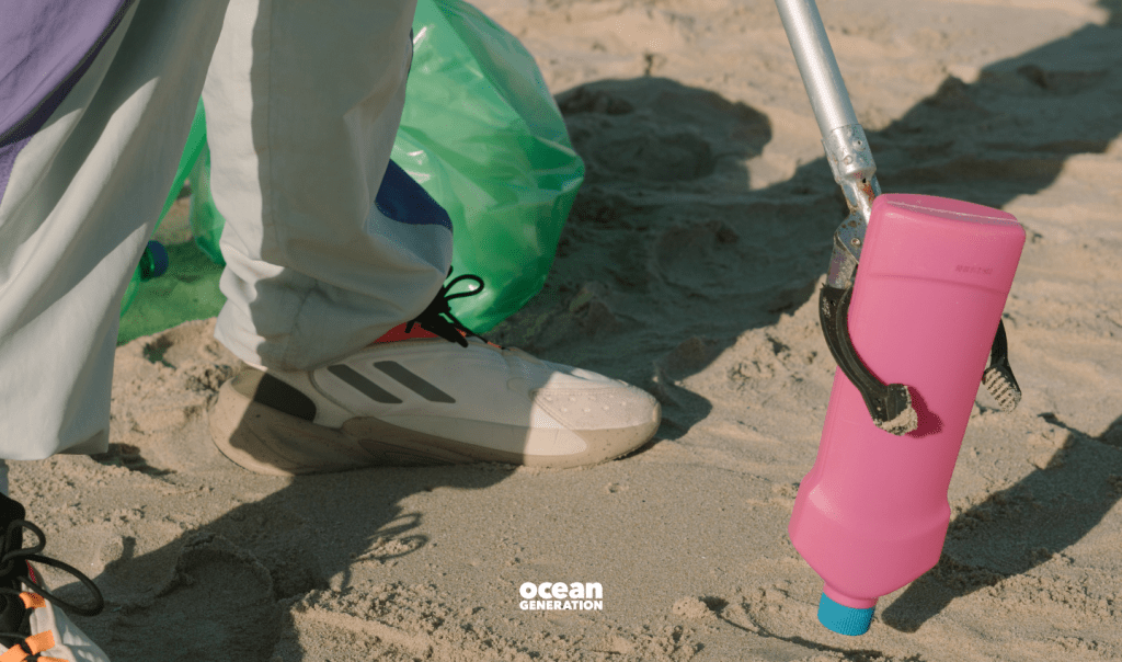 Scene from a beach clean. White tennis shoes of a litter picker are visible. The beach cleaner is picking up a pink plastic bottle off the beach. In the background, a plastic trash bag can be seen. Ocean Generation shares the history of plastic pollution in this blog.