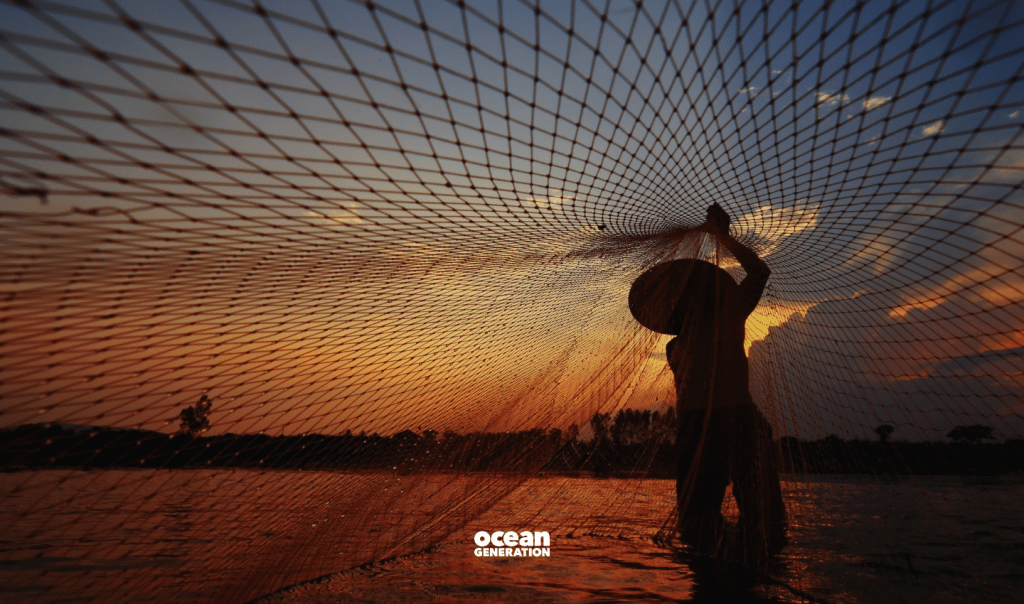 A fisherman, standing knee deep in the Ocean, is holding up a fishing net. It is sunset and only the outline of the fisherman and his hat can be seen against the yellow sky. In this blog, Ocean Generation is sharing the negative impact of resource extraction on the Ocean.