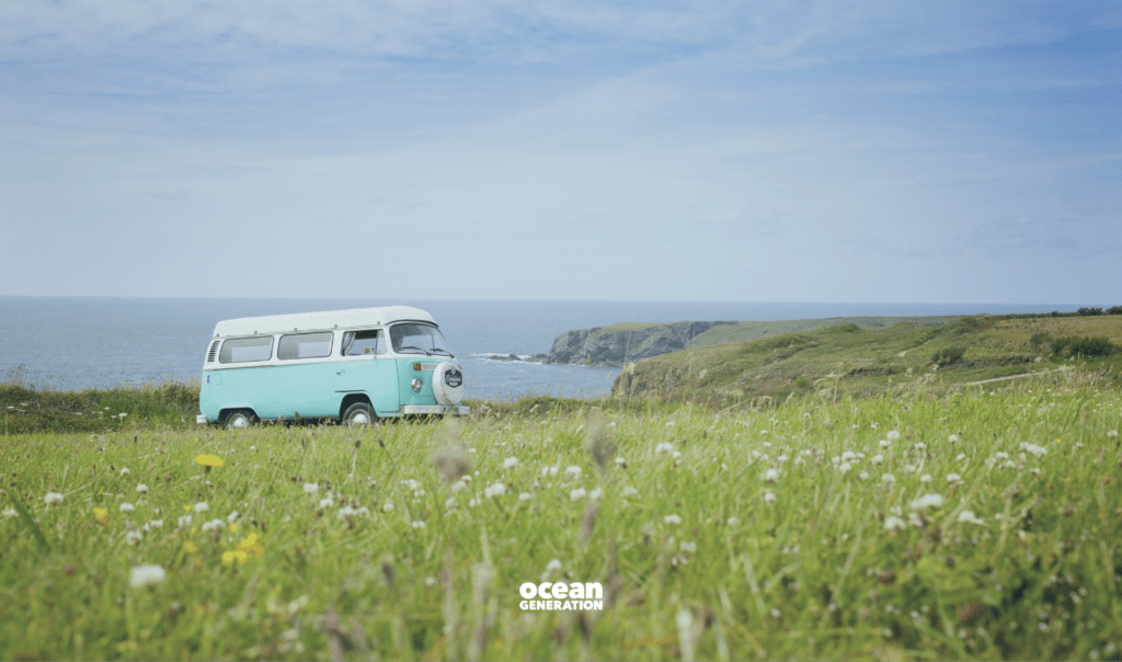 Teal travel van parked against the backdrop of a coastal road. There's a blue sky, a stretch of Ocean, and lush wild grass with a few flowers. Shared by Ocean Generation in an article about interesting travel facts with an environmental lens.
