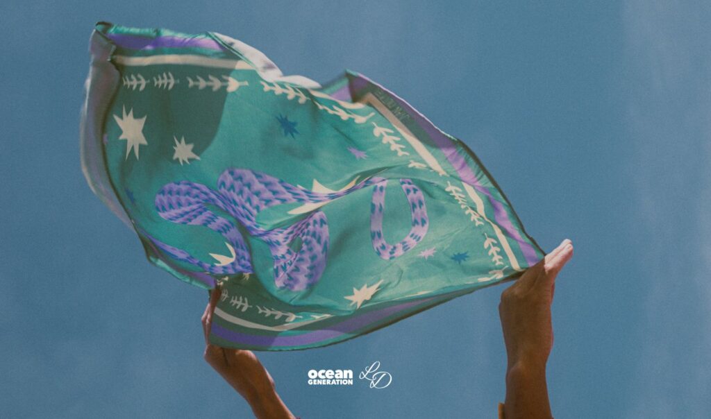 A sustainably made scarf rustles in the wind. It's being held up, by a set of hands. In the background is a blue sky. Shared by Ocean Generation: Experts in Ocean Health.